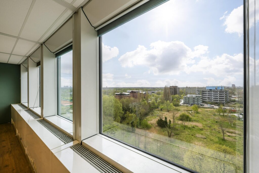 View from an office window overlooking the Laan van Kronenburg area with green spaces and buildings under a partly cloudy sky.